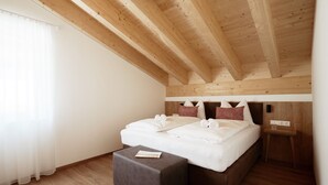 Un dormitorio con techo de madera, una cama con ropa de cama blanca, un reposapiés gris y una mesita de noche.