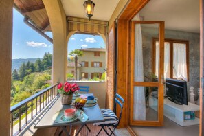 Ein Balkon mit Tisch für zwei Personen, einem Fernseher und Blick auf ein Haus und Hügel.