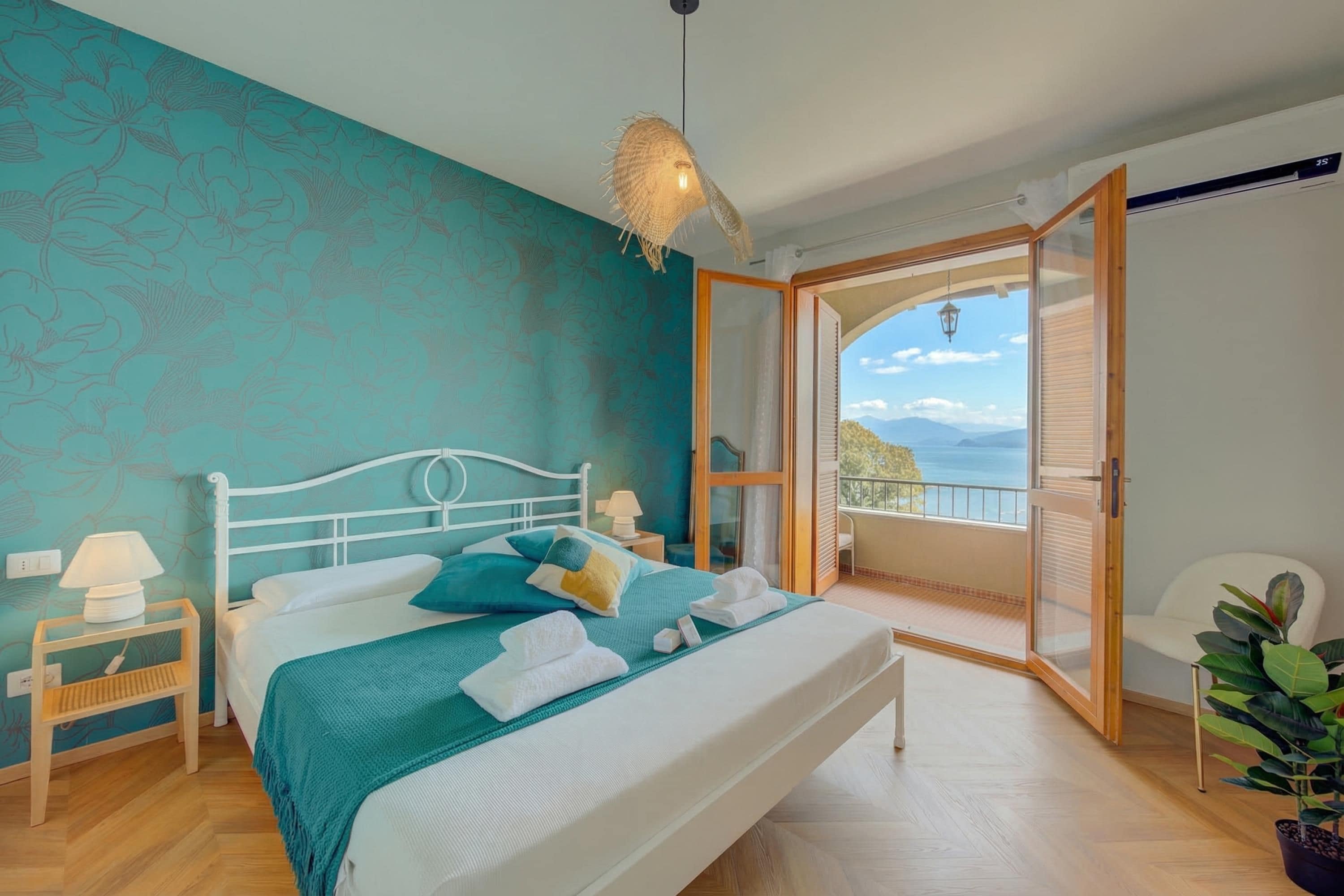 A bedroom with a bed, a nightstand, a lamp, a potted plant, and a view of the outdoors.