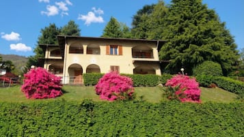 Un bâtiment à deux étages, doté d’une façade symétrique, de nombreux balcons et d’un jardin bien entretenu agrémenté d’arbustes à fleurs roses.
