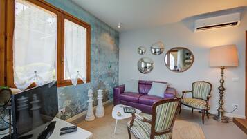 Un salon avec un canapé violet, un fauteuil à rayures, une table basse et un téléviseur. On peut voir des pièces d’échecs sur la table, et une lampe se trouve sur la droite.