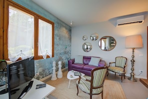 Un salón con un sofá morado, un sillón a rayas, una mesa de centro y un televisor. Hay piezas de ajedrez sobre la mesa, y una lámpara está a la derecha.