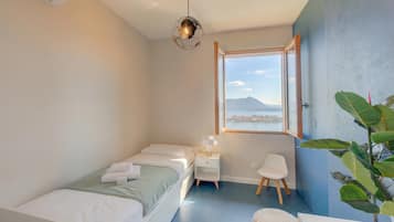 A modern bedroom with a bed, bedside table, chair, and a large window offering a view of the sea and mountains.