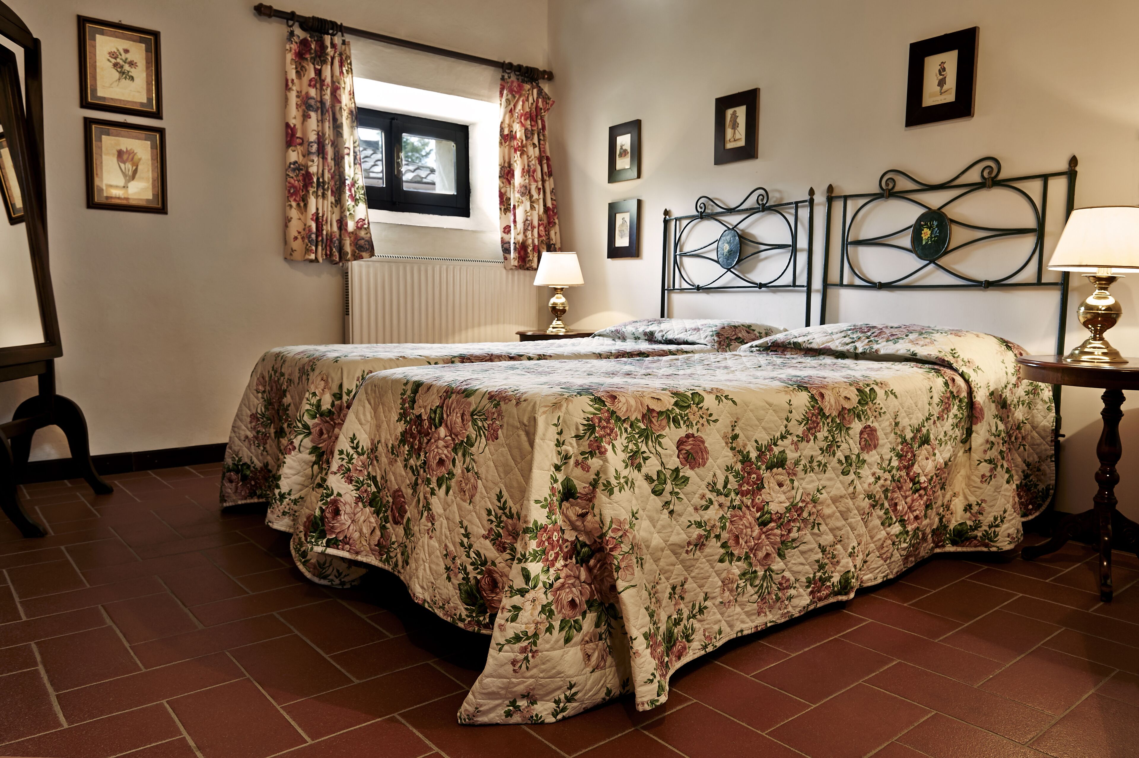 A bedroom with a floral bedspread, a window with curtains, a bedside table with a lamp, and framed pictures on the wall.
