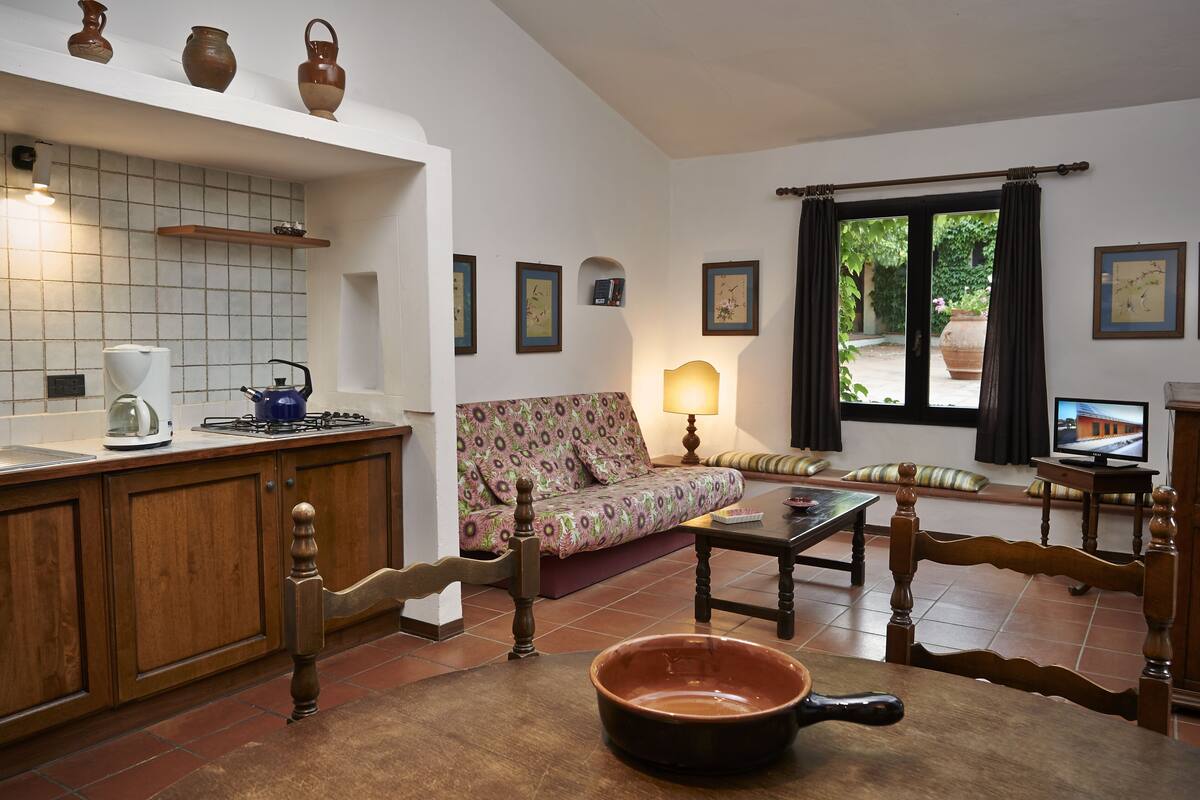 Eine Küche mit Herd, Wasserkocher und Kaffeemaschine, ein Essbereich mit Tisch und Stühlen, ein Sofa und ein Fenster mit Vorhängen.