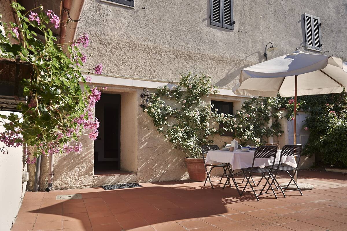 Eine Terrasse im Freien mit Tisch und Stühlen, einem Blumentopf und einem Gebäude mit einer strukturierten Wand und Fensterläden.