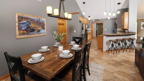 A dining area with a wooden table set for four, surrounded by chairs. There is a kitchen in the background with a bar area and stools. - 3br, sleeps 10, Gatlinburg, all the amenities! (Gatlinburg)