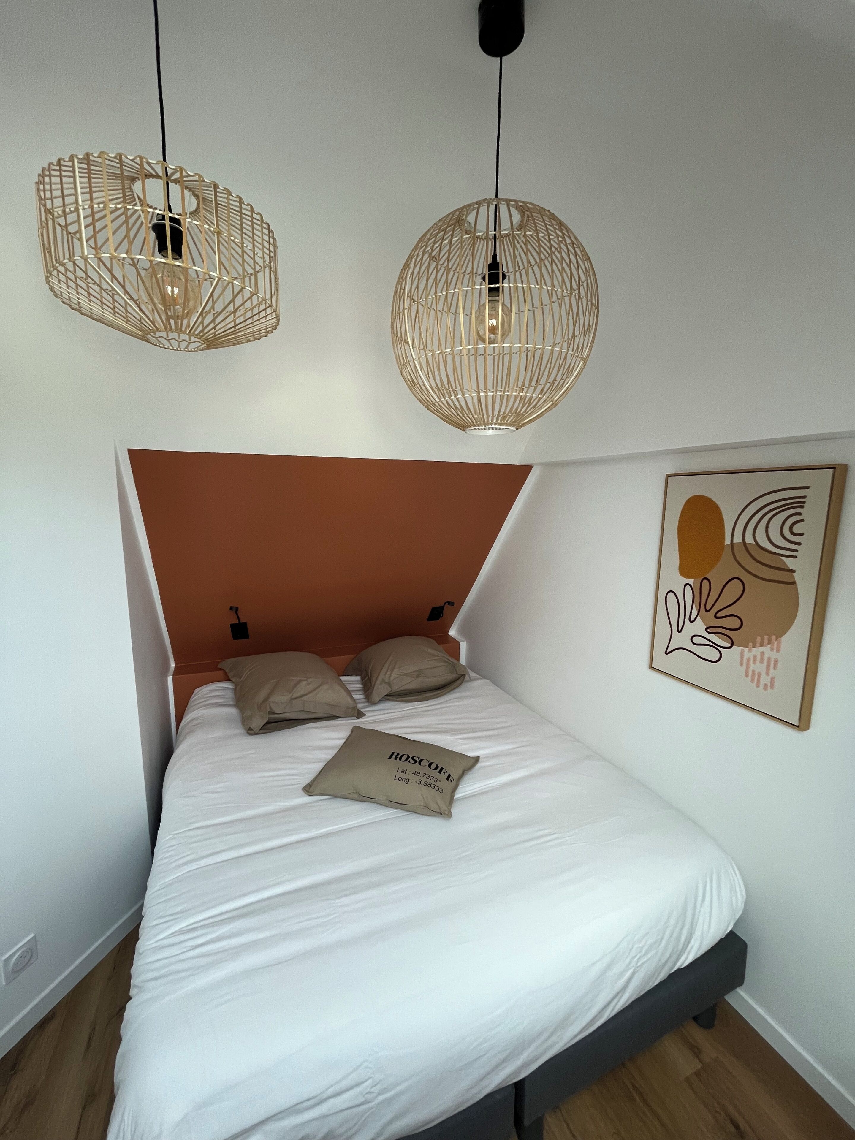 A bedroom with a bed, two hanging light fixtures, a headboard, a pillow, and a wall art piece.