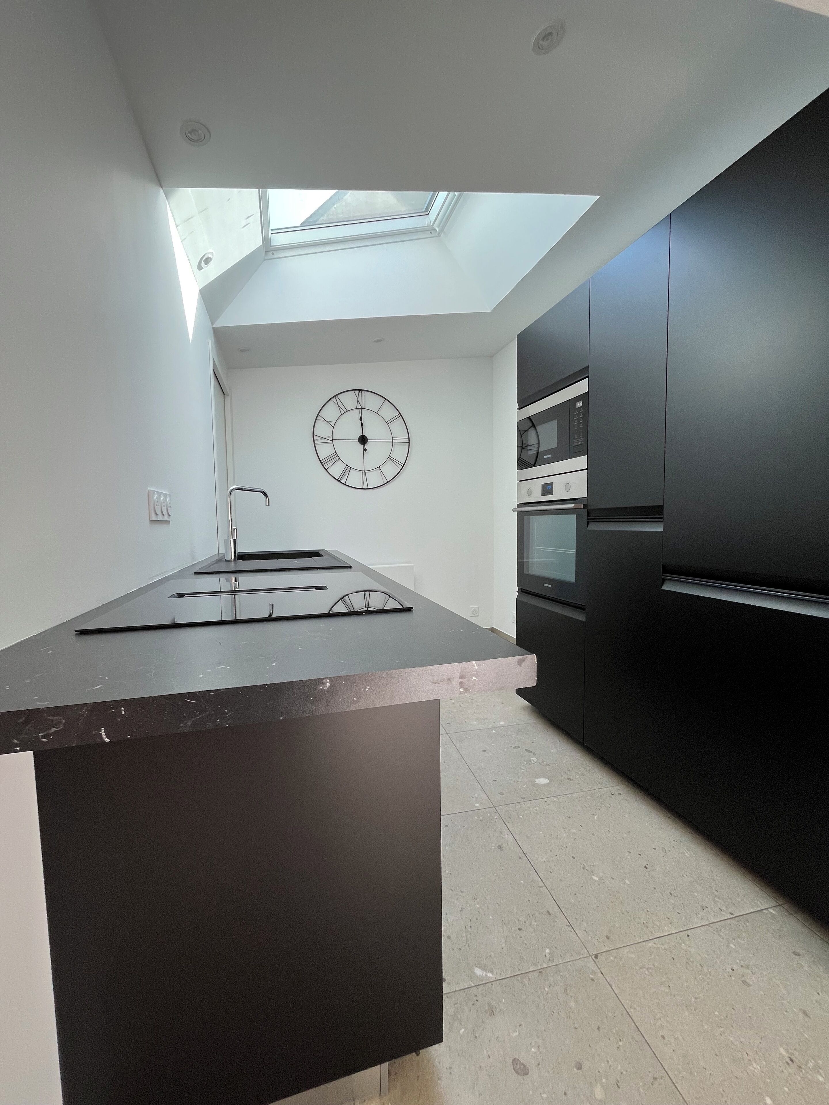 A modern kitchen with a skylight, a large clock on the wall, and dark cabinetry.