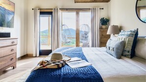 Ein Schlafzimmer mit einem großen Bett, einer Holzkommode, einem Hängekorbstuhl und Blick ins Freie.