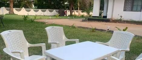 Uma mesa de plástico branca e quatro cadeiras dispostas numa área gramada, com um edifício branco ao fundo.