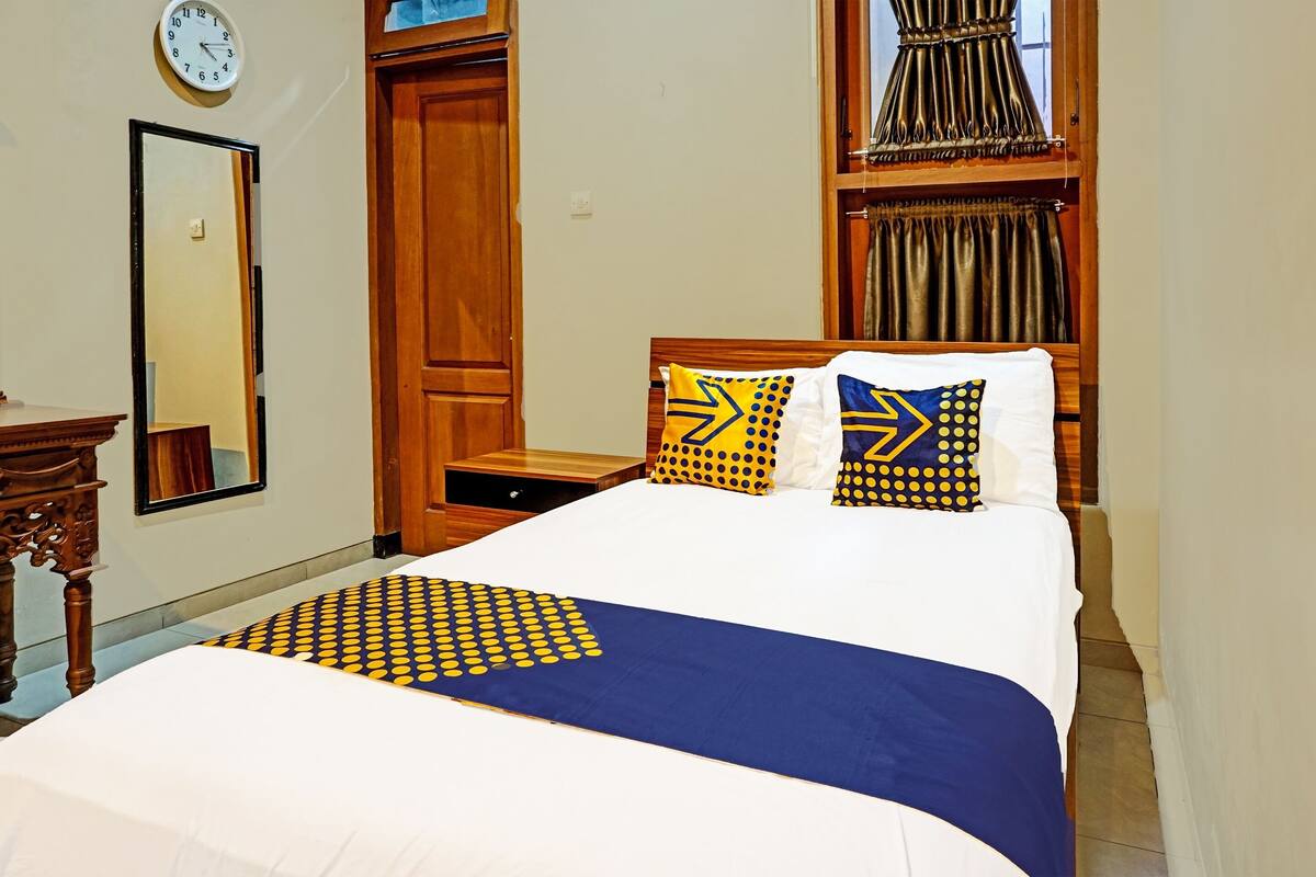 Un lit bien fait, recouvert d’une couvre-lit à motifs bleus et jaunes, deux oreillers aux motifs géométriques, une table de chevet en bois, une armoire en bois, un miroir et une horloge au mur.