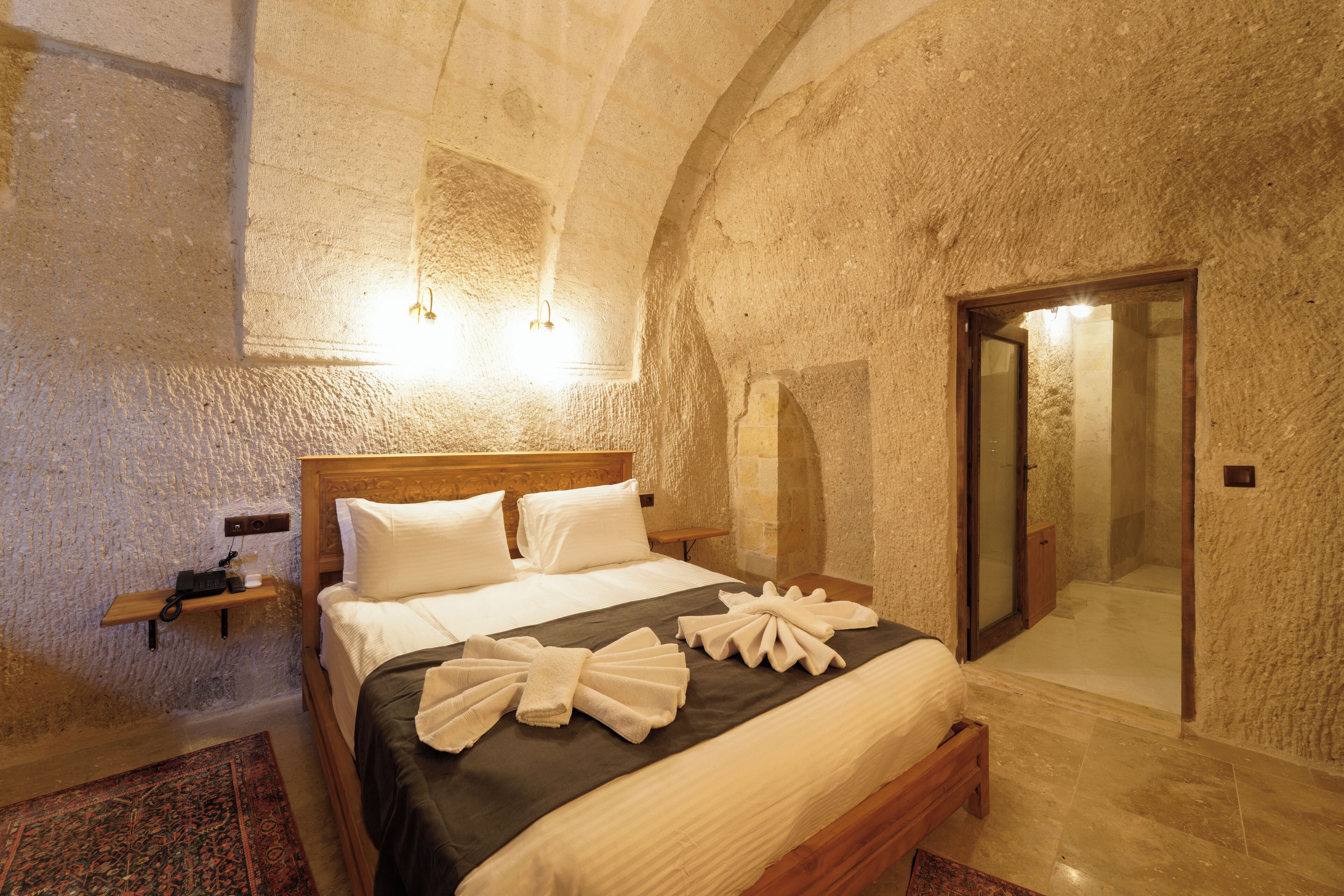 A hotel room with a large bed, a wooden headboard, a nightstand, a bedside table, a wall-mounted light, and a shower area.