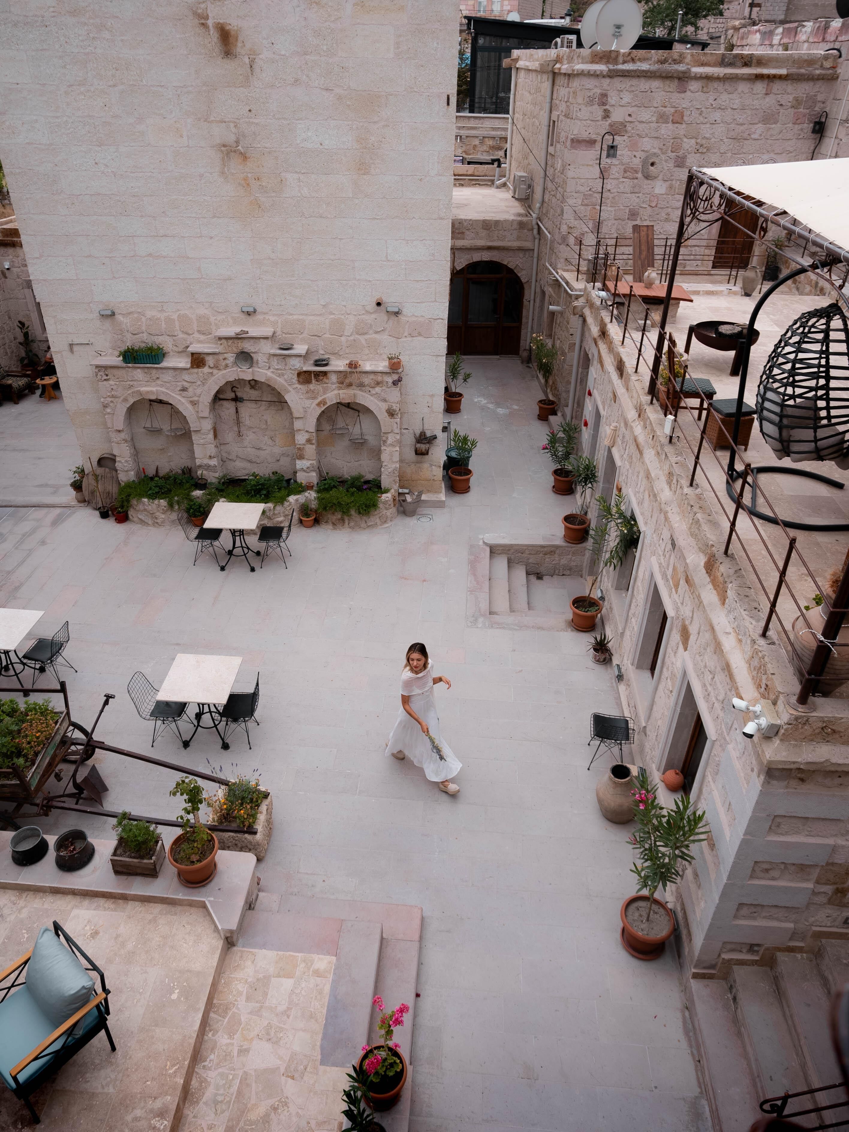 An outdoor courtyard with stone walls, potted plants, outdoor seating, and a person walking.