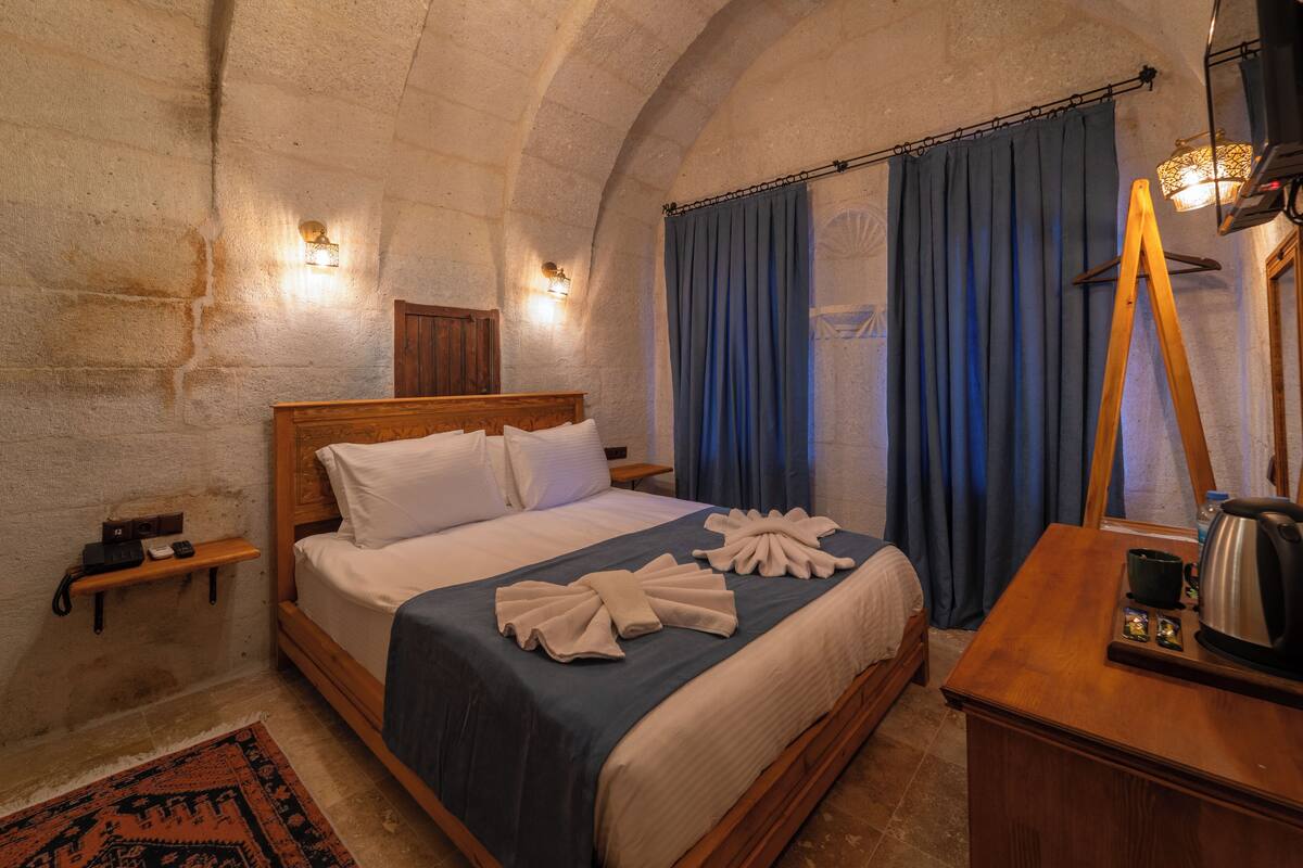 A room with a wooden bed, a nightstand, a desk with a kettle, and a blue curtain.