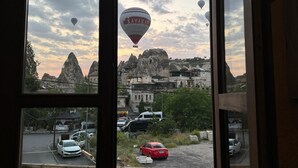 Une vue à travers une fenêtre sur une rue où l’on aperçoit plusieurs montgolfières dans le ciel, une voiture rouge sur la route et des bâtiments en arrière-plan.