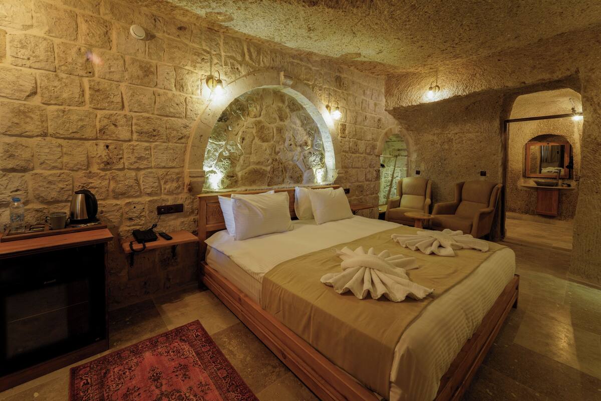 A stone-walled bedroom with a bed, a wooden nightstand, a red rug, and a mirror above the bed.