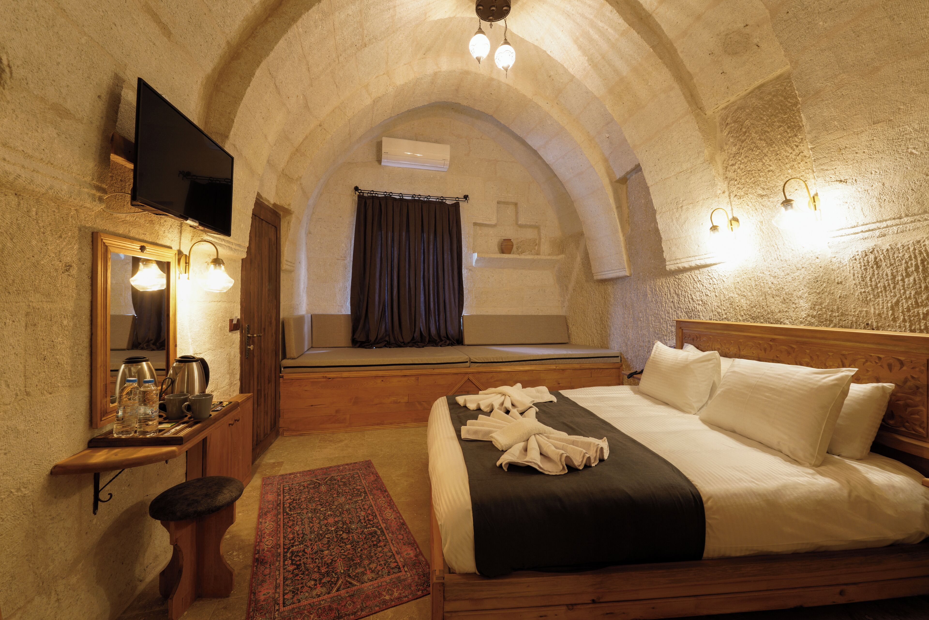A bedroom with a wooden bed, a nightstand with a mirror, a television, and a stone wall.