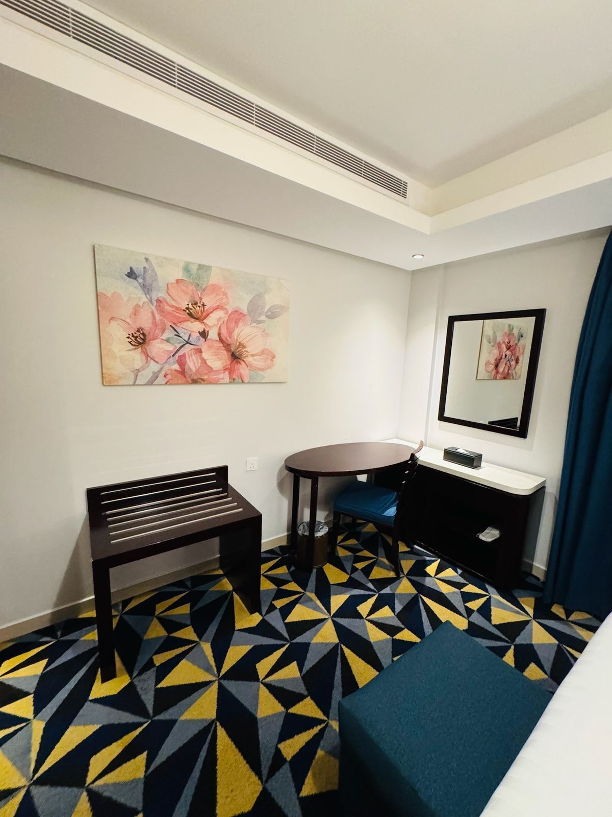 Una habitación de hotel con una alfombra de diseño geométrico, un banco, una mesita, un escritorio con un espejo y un cuadro en la pared.