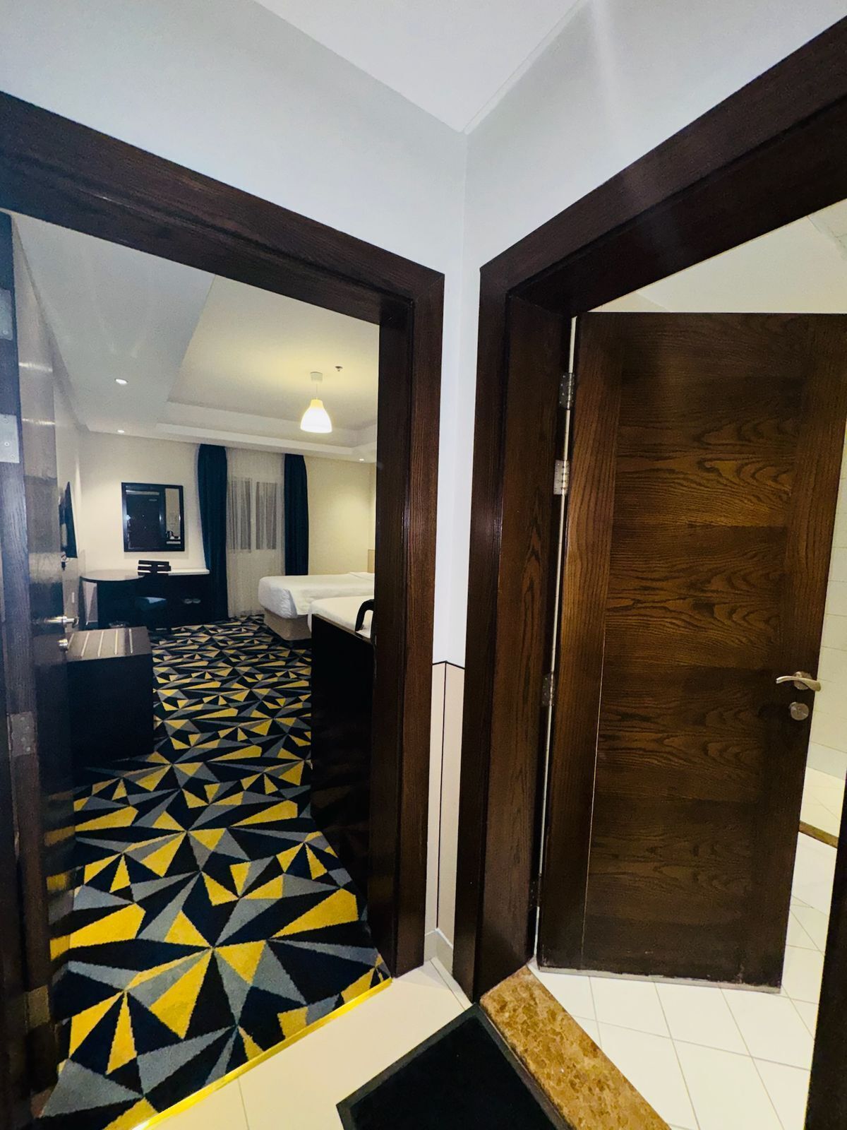 Un pasillo de hotel con suelo de diseño geométrico, una cama, un baño y una puerta de madera.