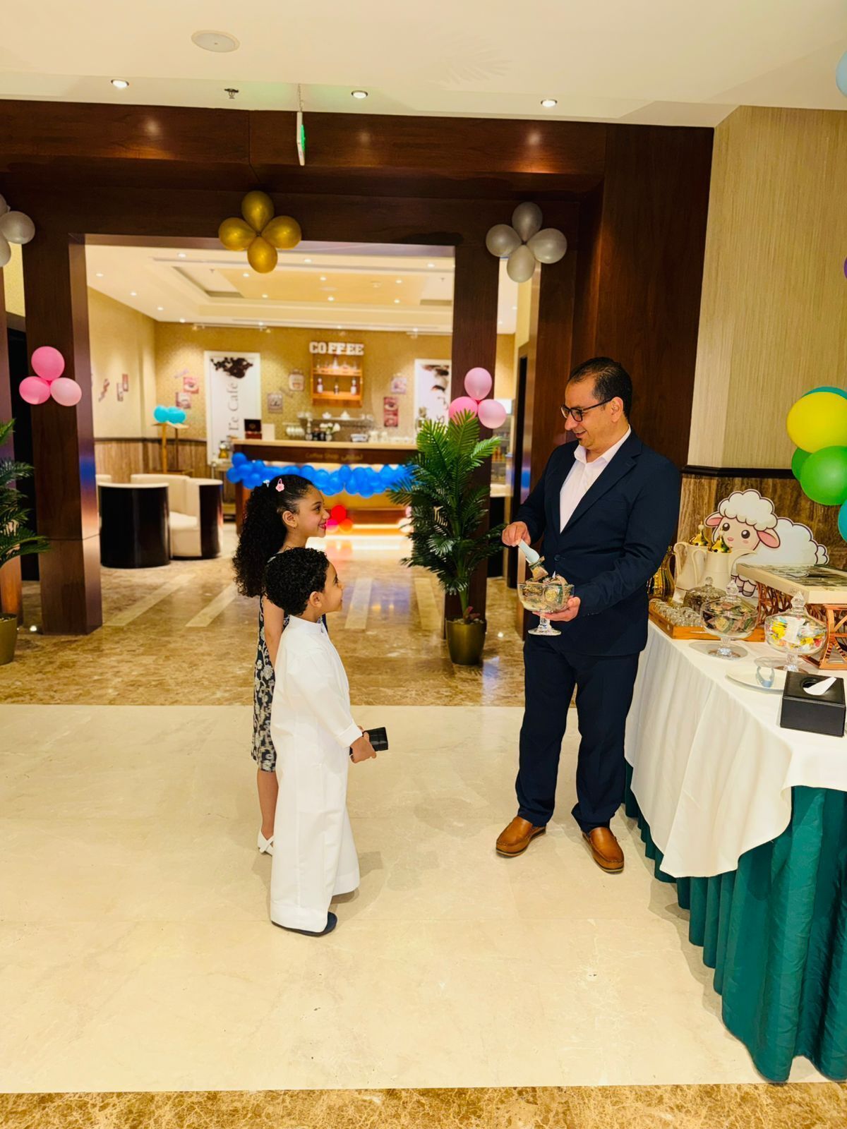 Un hombre con traje sostiene un vaso, de pie junto a una niña con vestido blanco, en una habitación con una cafetería al fondo y globos.