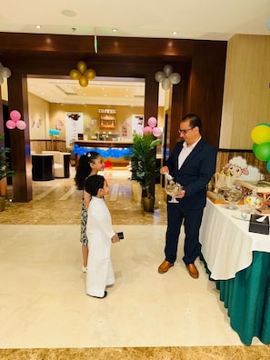 Un hombre con traje sostiene un vaso, de pie junto a una niña con vestido blanco, en una habitación con una cafetería al fondo y globos.