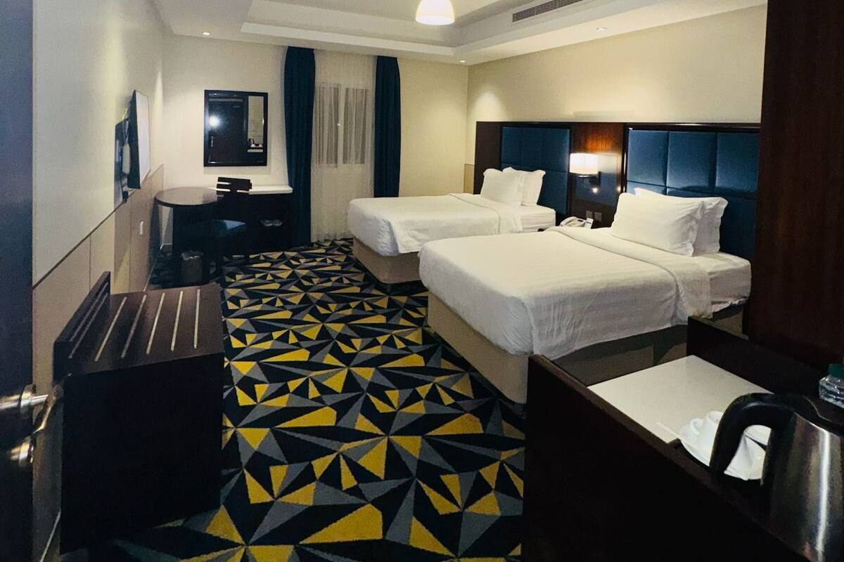 Quarto de hotel com duas camas, uma escrivaninha, uma cadeira, uma televisão e um chão com padrão.