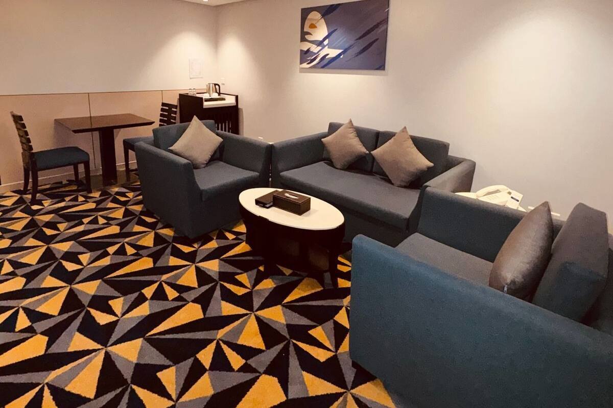 Uma sala de estar moderna com carpete estampado em padrão geométrico, sofá modular, pequena mesa de centro redonda e mesa de jantar com cadeiras.