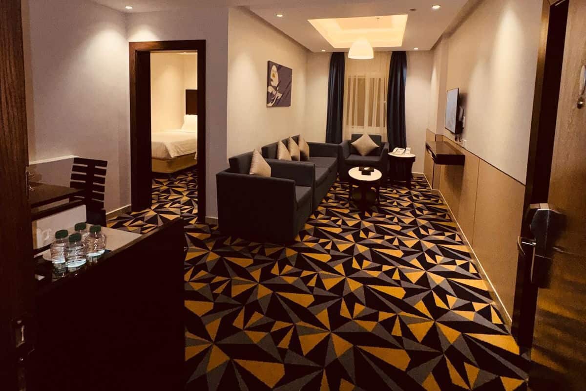 Quarto de hotel com carpete estampado em padrão geométrico, um sofá, uma pequena mesa e uma cama.