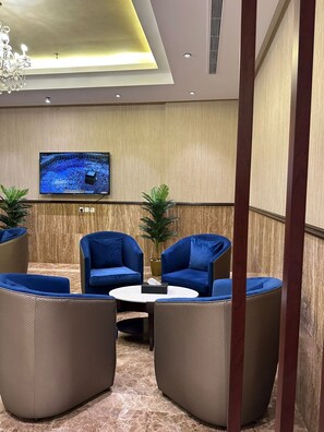 A modern lounge area with blue armchairs, a round table, and a television displaying a news broadcast. - Ocean View Al Zahra (Jeddah)