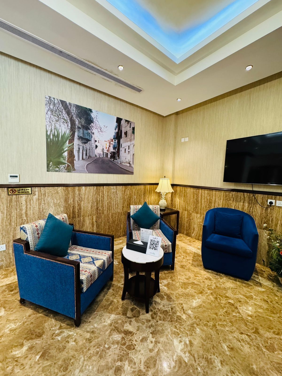 A hotel room with a marble floor, two sofas with patterned upholstery, a small table with a lamp, a flat-screen TV, and a large framed picture on the wall.