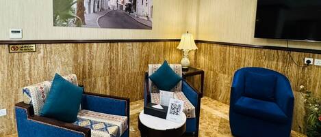 A hotel room with a marble floor, two sofas with patterned upholstery, a small table with a lamp, a flat-screen TV, and a large framed picture on the wall.