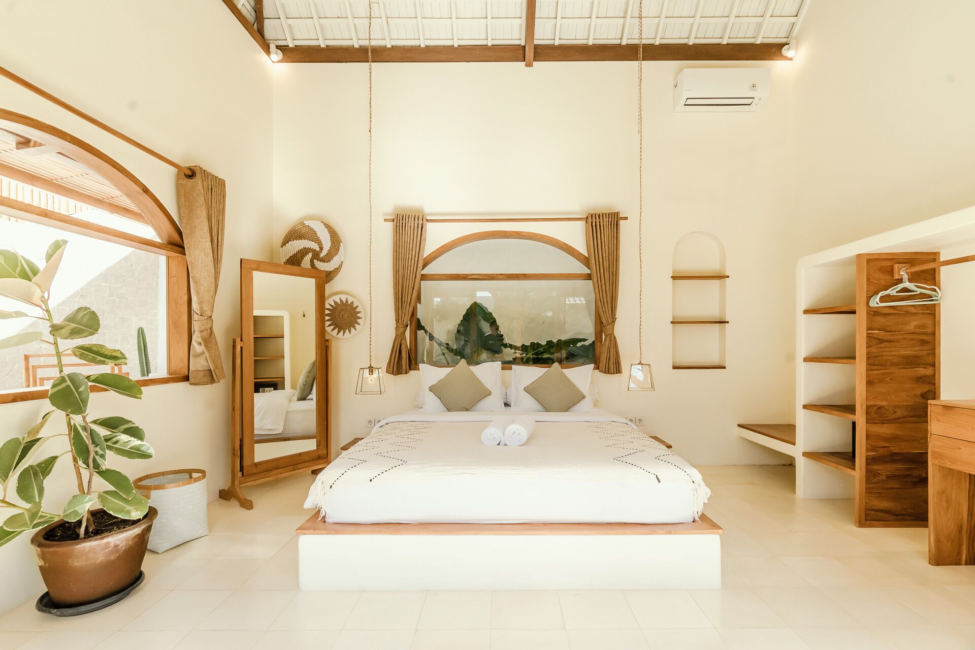 A bedroom with a large bed, a wooden wardrobe, a mirror, and a potted plant.