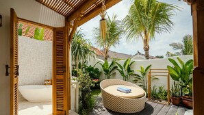 A private outdoor bathroom with a bathtub, wooden door, and a round seating area with a cushion. - Canggutopia by Alfred in Bali (Canggu)