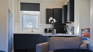 A modern kitchen with dark cabinetry, a window with a view, and a dining area with a table and chairs. - The White City Apartment (Oban)