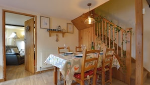 Ein Essbereich mit einem Holztisch, gedeckt für zwei Personen, umgeben von Stühlen mit roten Kissen. Im Hintergrund ist eine Holztreppe mit Grünpflanzen zu sehen.