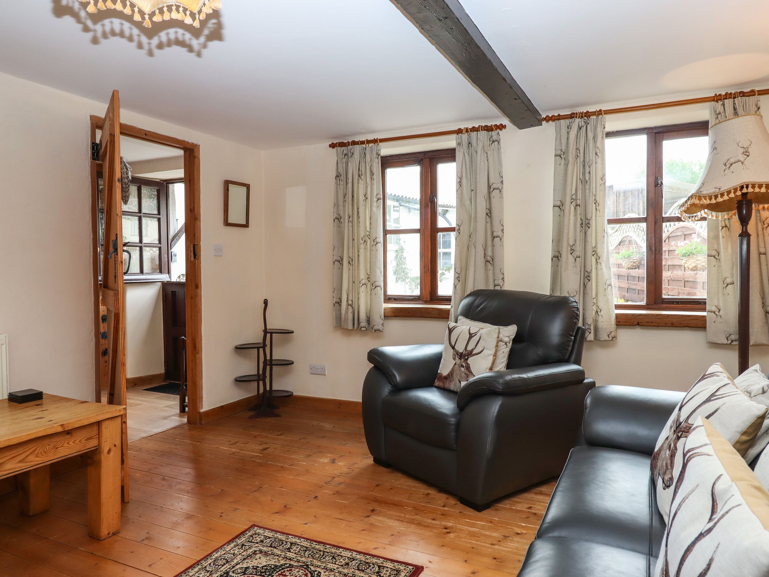 A living room with a wooden floor, a black leather sofa, a wooden coffee table, and a patterned rug. There are two windows with curtains, a chandelier, and a door leading to another room.