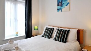A hotel room with a bed, a nightstand, a lamp, a window with curtains, and a painting on the wall. - Admiral's Quarter Apartment 5 (Weymouth)