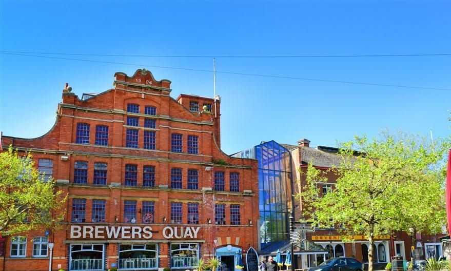 Un edificio de ladrillo rojo con un letrero que dice "BREWERS QUAY" y una estructura con fachada de vidrio adyacente.
