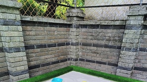 Un espace de jeux carré recouvert de gazon synthétique, entouré d’un mur de briques et d’une clôture en treillis métallique.