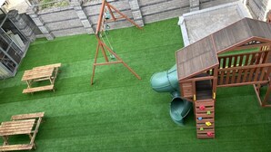Un jardin avec une pelouse artificielle, une balançoire en bois, un toboggan et une cabane de jeux.