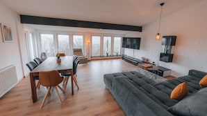 A modern living room with a dining area, grey sofa, and a TV mounted on the wall. - RhönKristall modern terrace house with a far-reaching view (Bischofsheim in der Rhön)