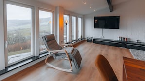 A spacious living room with large windows, a modern sofa, a wooden coffee table, and a flat-screen TV mounted on the wall. - RhönKristall modern terrace house with a far-reaching view (Bischofsheim in der Rhön)
