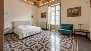 Une chambre à coucher avec un grand lit, un fauteuil bleu, une petite table et une fenêtre avec des rideaux.