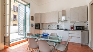 Une cuisine moderne avec une table à manger en verre, des chaises de couleur beige et une vue sur la rue grâce à une porte ouverte.