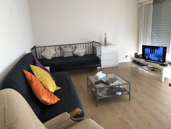 Una sala de estar con un sofá negro, una mesa de centro de vidrio, un televisor y un mueble blanco.