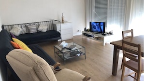 Ein Wohnzimmer mit einer schwarzen Couch, einem Glastisch, einem Holzesstisch und einem Fernseher auf einem weißen Schrank.