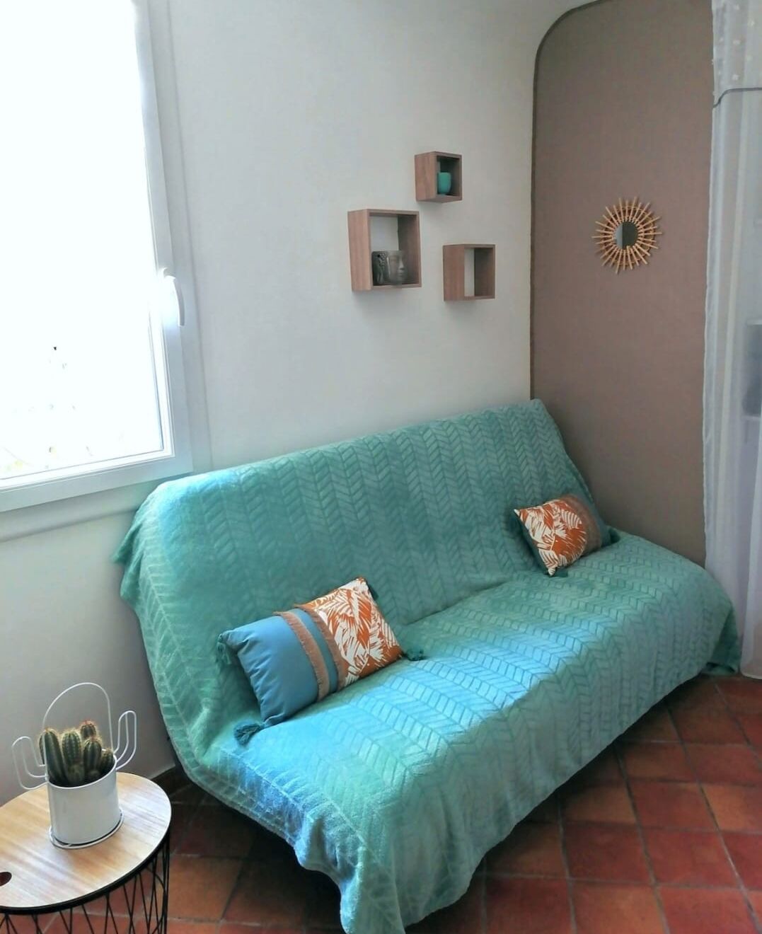 A teal sofa with patterned cushions, a small cactus on a round table, and a wall-mounted decorative item.