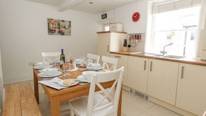 A dining area with a wooden table set for four, white chairs, a kitchen with a sink and cabinets, and a clock on the wall. - Harbour Tales (Weymouth)