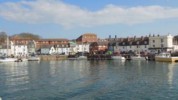 A harbor with boats docked, buildings along the waterfront, and a clear sky. - Harbour Tales (Weymouth)